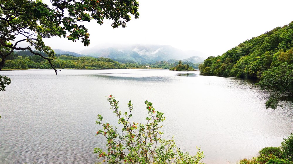 Serene lake with surrounding green hills and misty mountains in the background.
