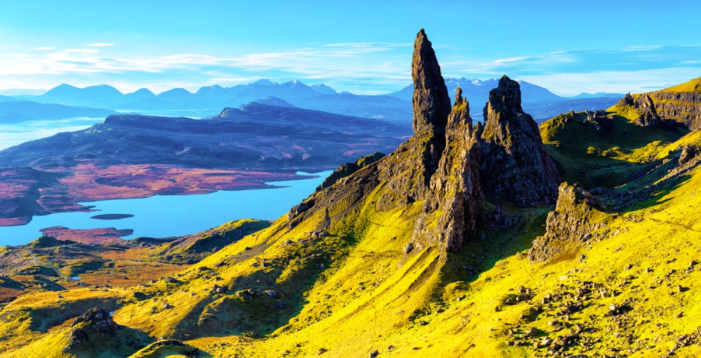 Mountainous landscape with rocky peaks, green slopes, and distant water view under a clear blue sky.