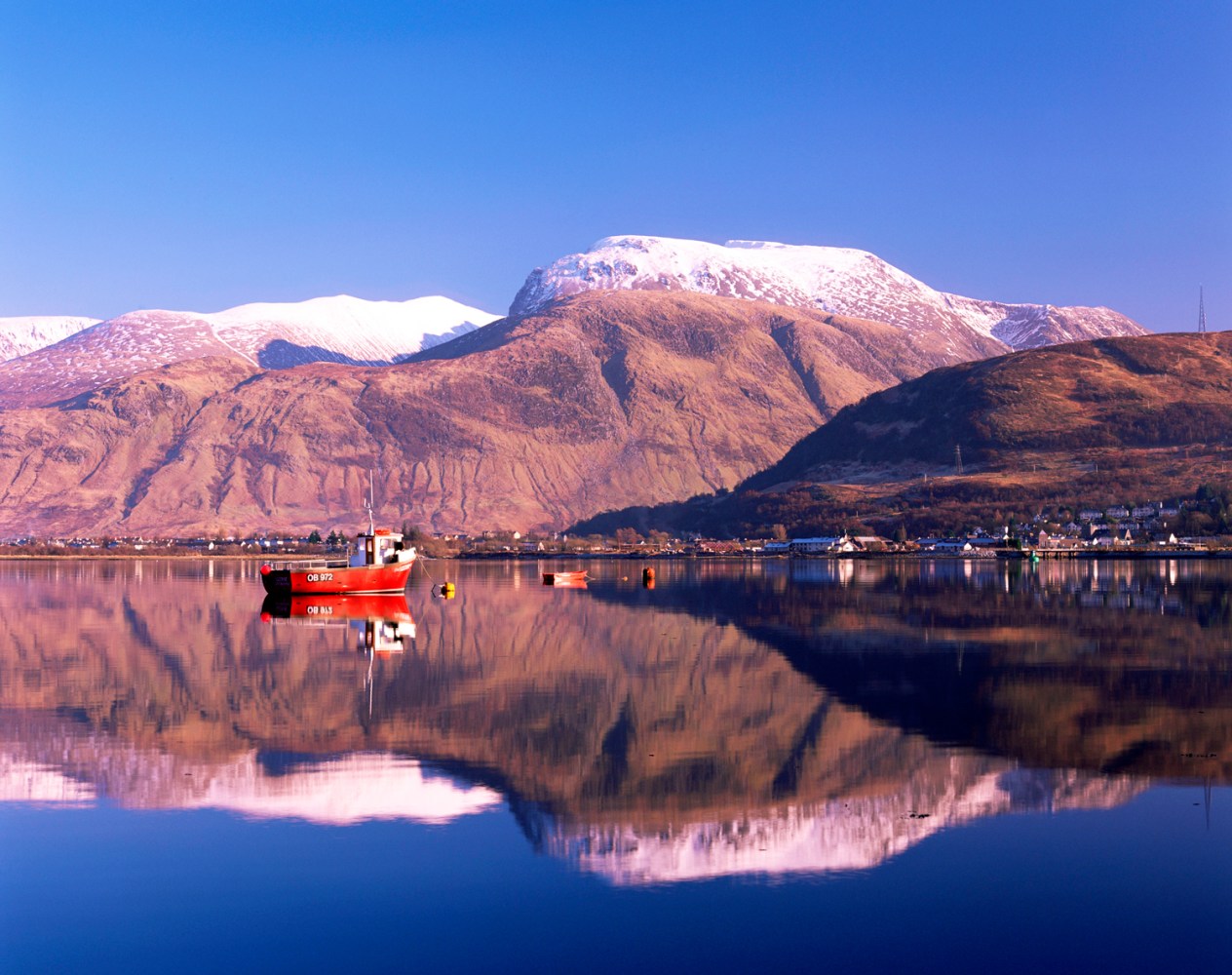 Snow-capped mountains and red boat reflected in calm lake.