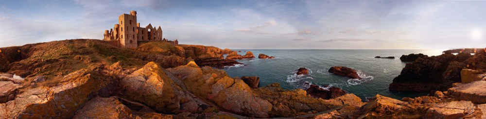 Panoramic view of a castle on a rocky coast with ocean waves and a sunset sky.