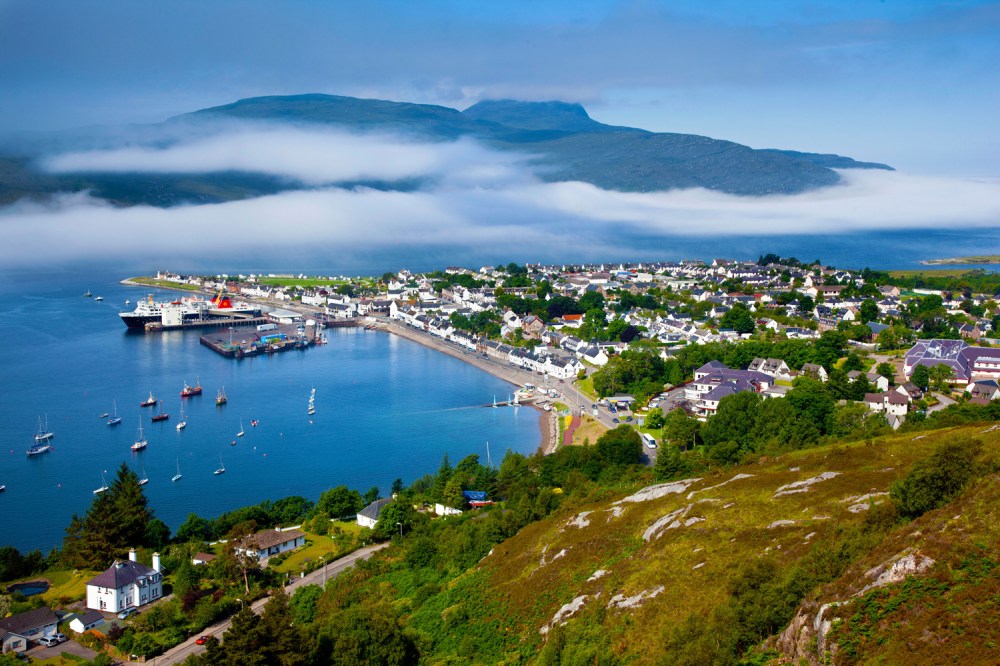 Coastal town with harbor, sailboats, houses, and distant mountains under a blue sky with clouds.