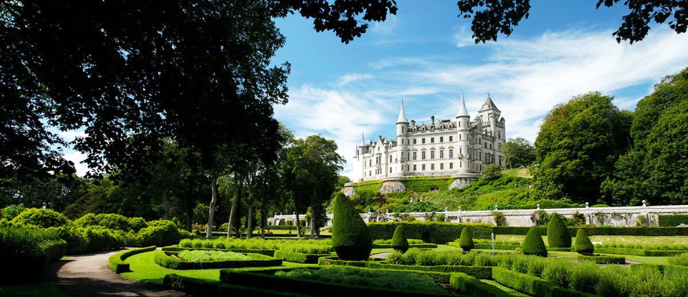 Elegant castle with spires surrounded by lush gardens and trees under a blue sky.
