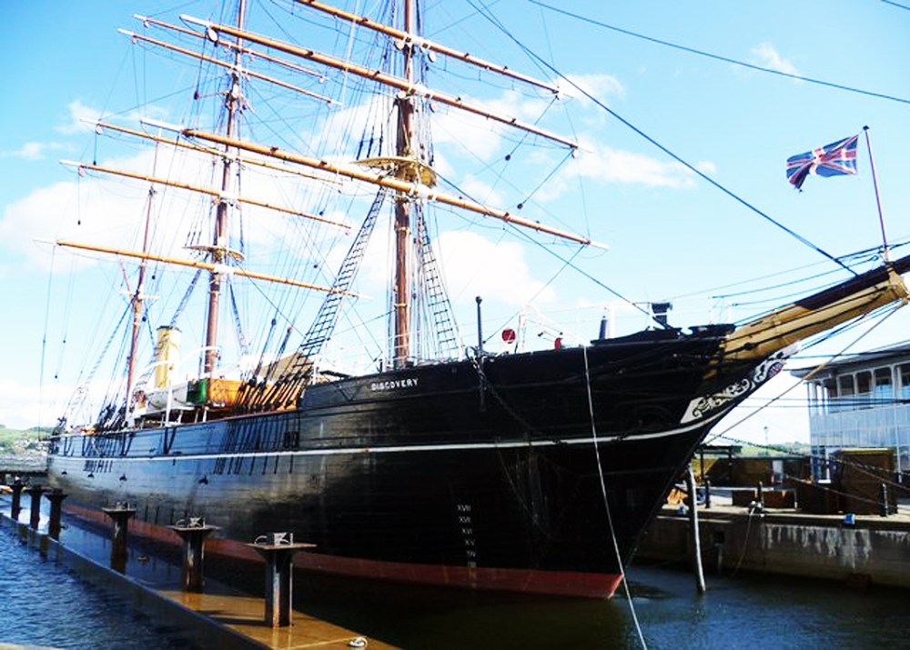 A large, historic tall ship with multiple masts docked, Union Jack flag visible at the stern.