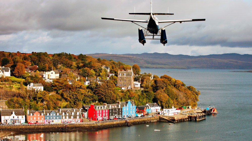 Seaplane flying over colorful coastal village with hillside and water view.