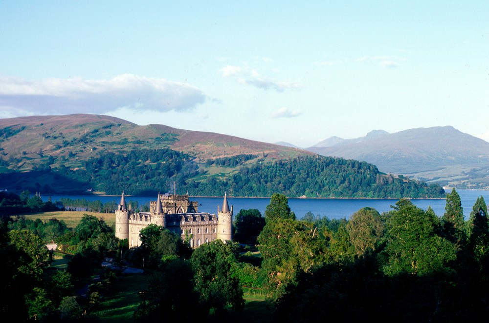 Castle near a lake, surrounded by trees and hills under a clear blue sky.