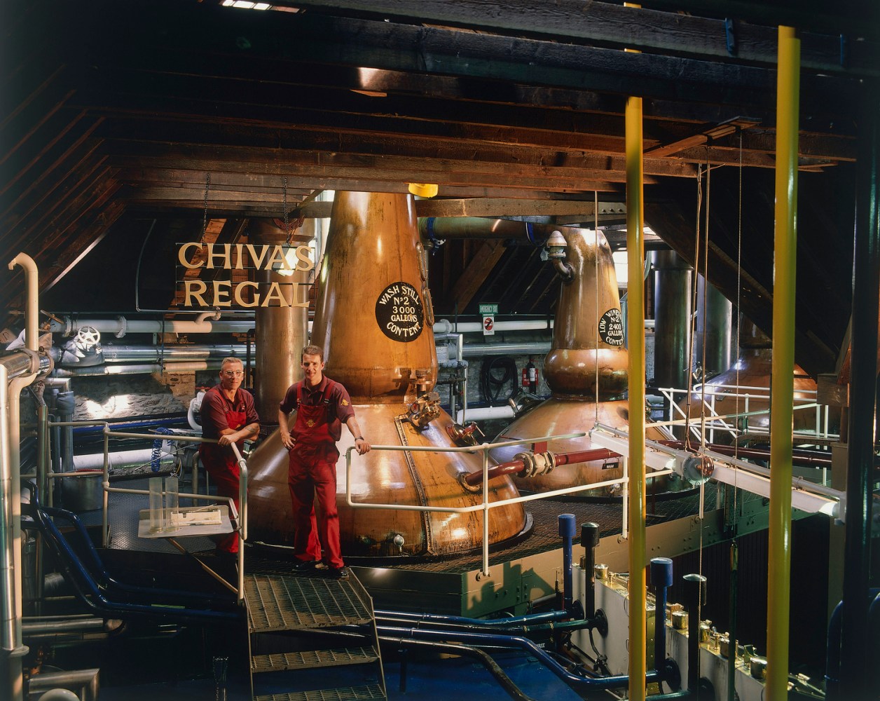 Two workers in a whiskey distillery with copper pot stills labeled 'Chivas Regal'.