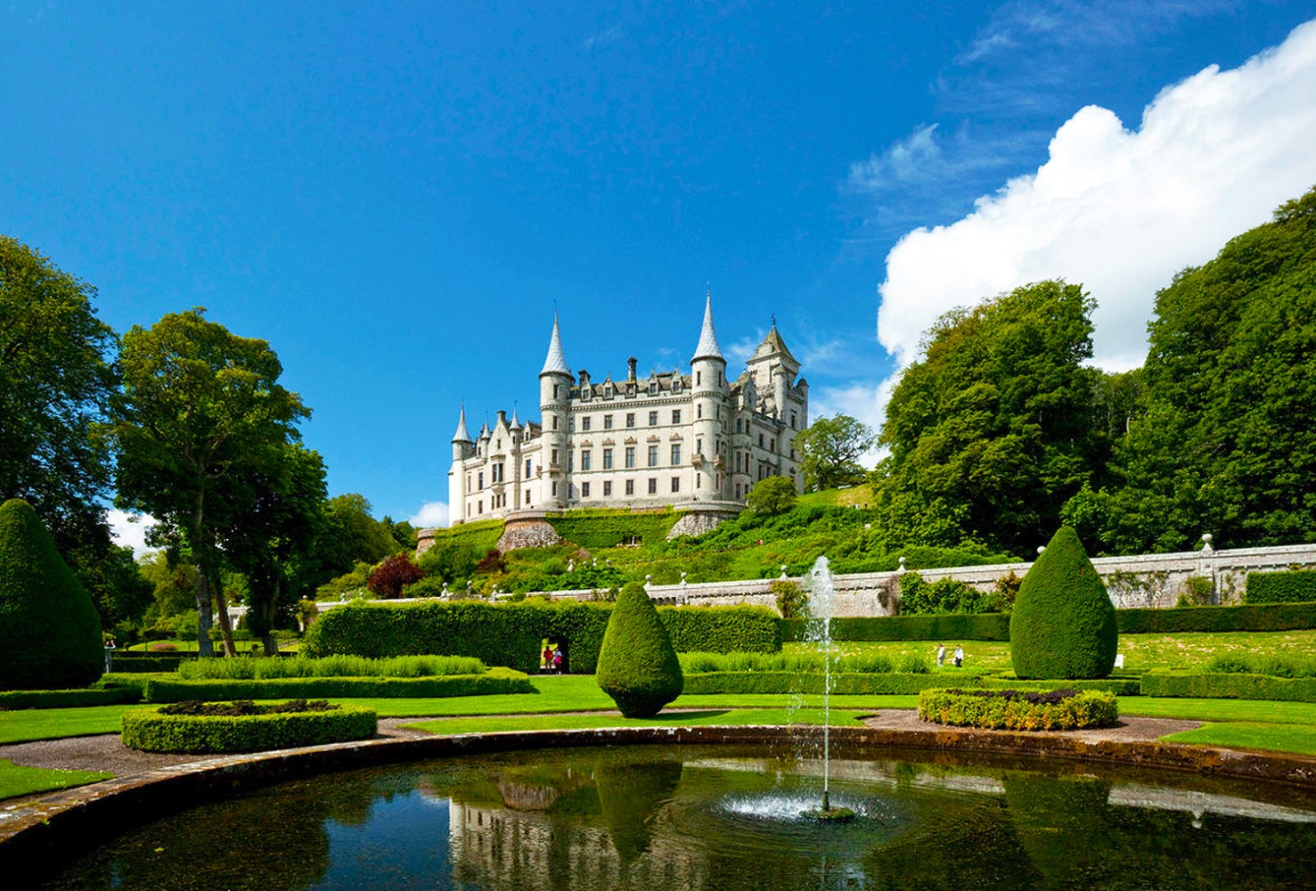 Large castle with towers, surrounded by lush gardens and a pond, under a clear blue sky.