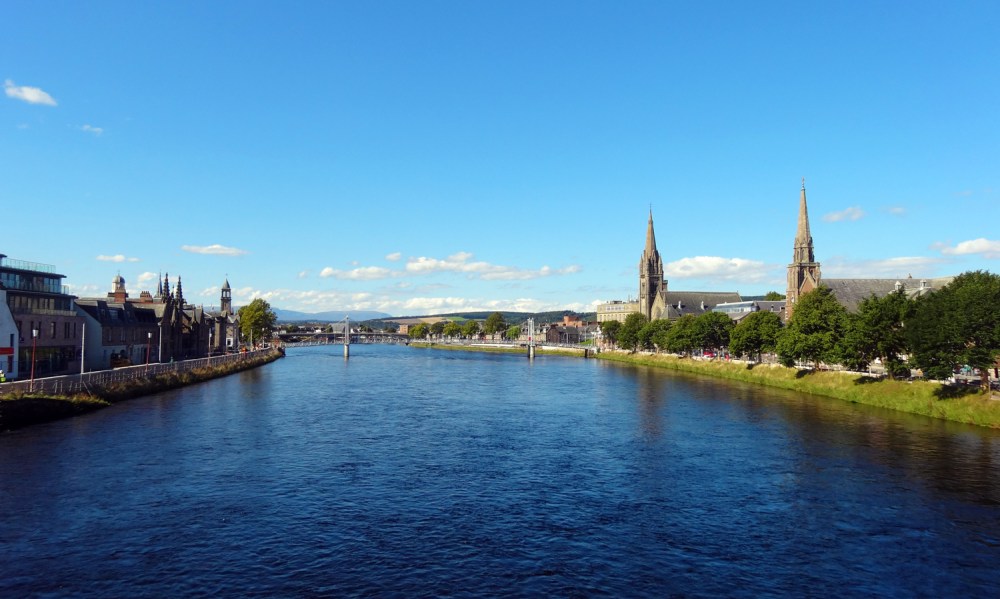 View of a river with city buildings and two church spires under a clear blue sky.