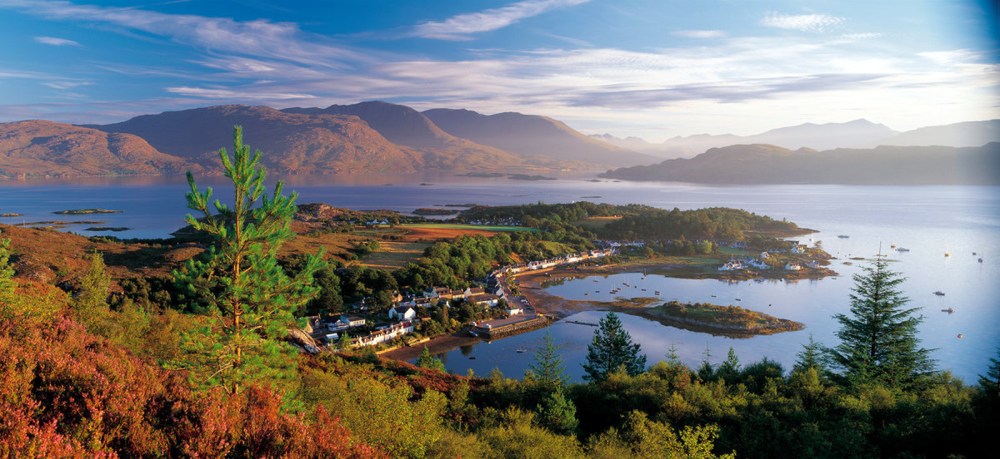 Scenic view of a coastal village with mountains and a bay, framed by trees and under a partly cloudy sky.