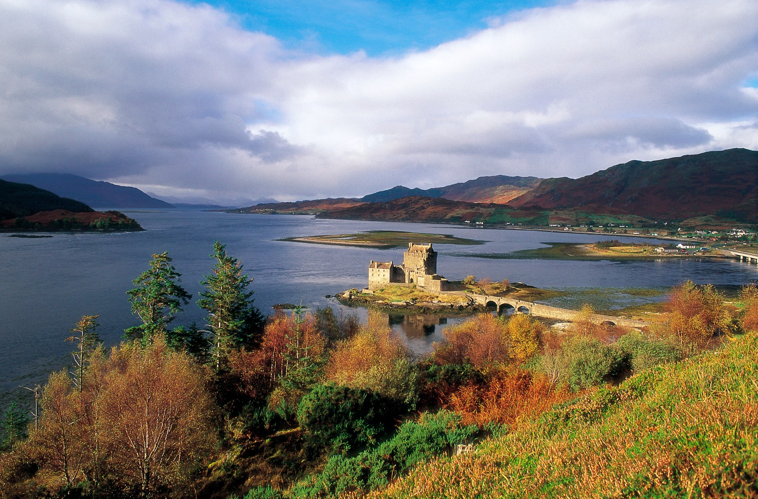 Castle on a small island with a bridge, surrounded by water and mountains.