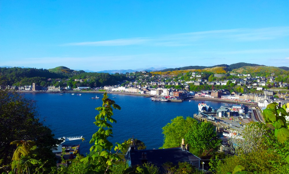 Coastal town with harbor, buildings, and hillside under clear blue sky.