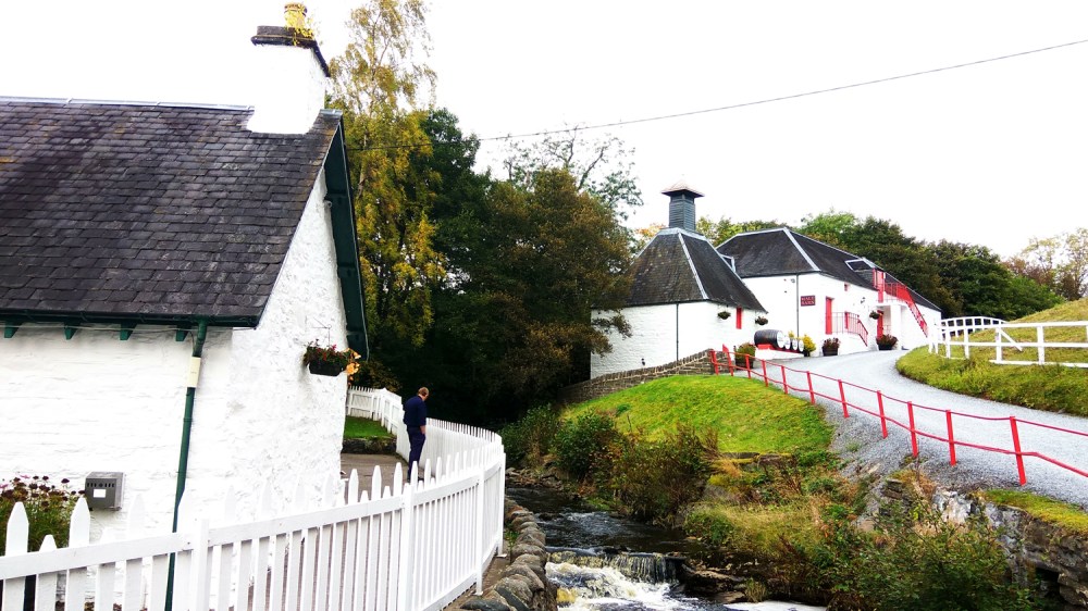 White cottages with chimneys, red doors by a stream and bridge, surrounded by greenery.