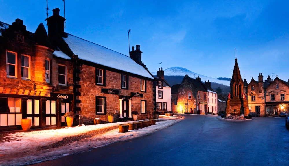 Snow-covered village street at dusk with lit buildings and a mountain backdrop.