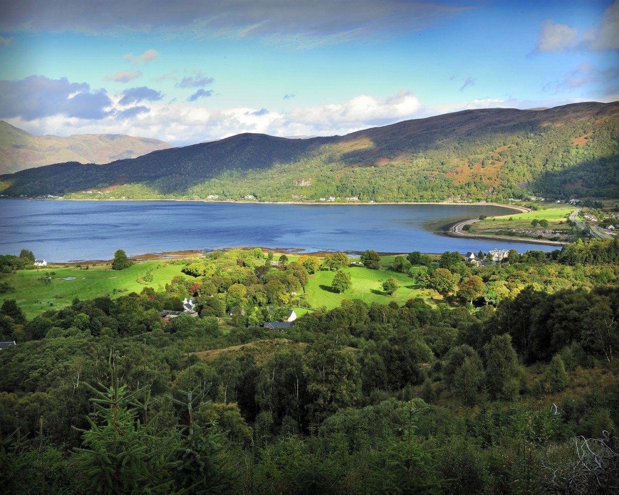 Scenic view of a lush green valley with hills, a lake, and a blue sky with clouds.