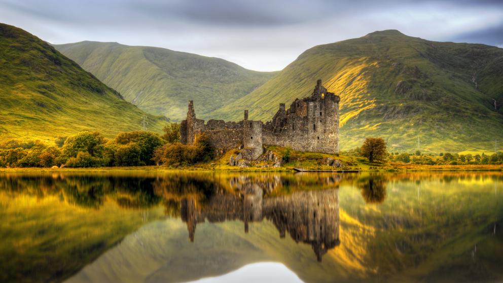 Ruinas de un castillo reflejadas en un lago tranquilo y cristalino, rodeado de exuberantes colinas verdes.