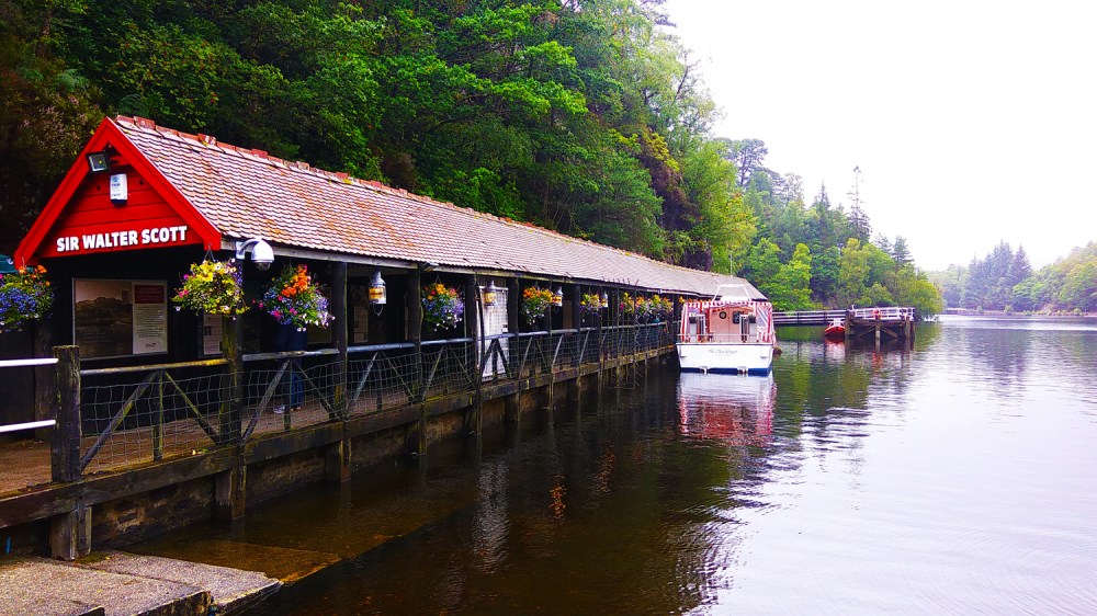 Boathouse with hanging plants by a lake in a wooded area.