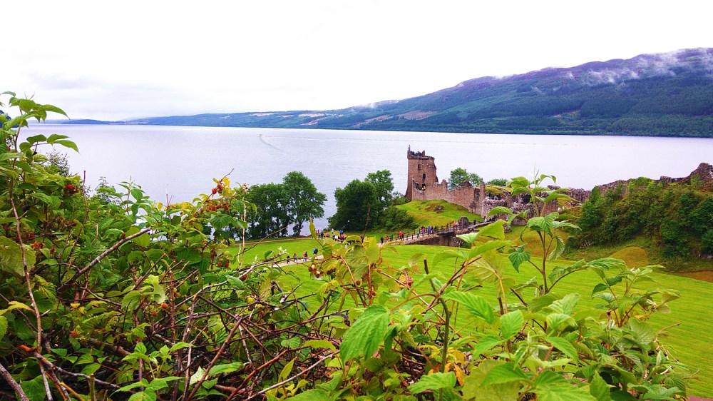Castle ruins beside a large lake with hills and trees in the background.