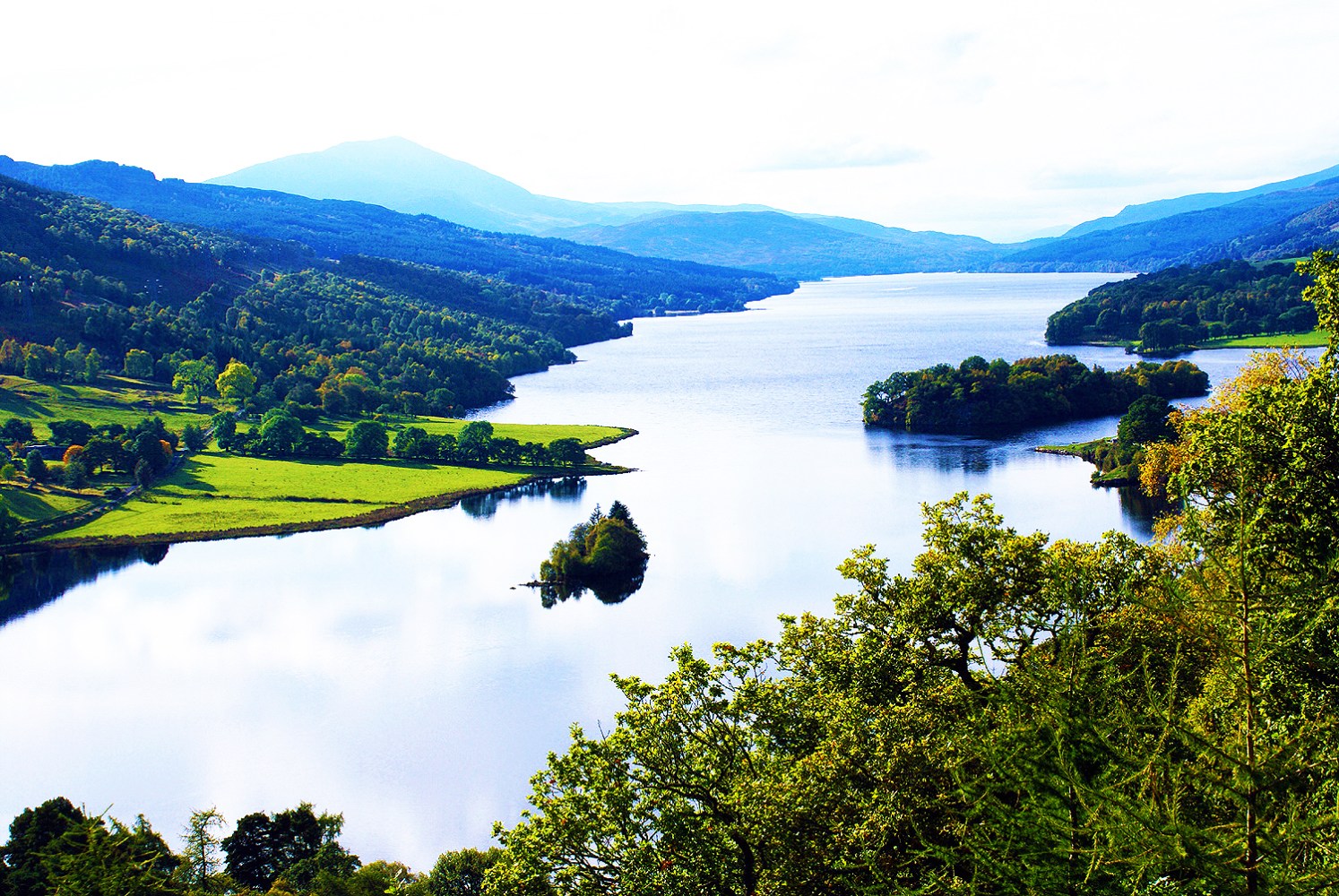 Impresionante vista panorámica del lago con árboles, pequeñas islas y colinas a lo lejos bajo un cielo despejado.
