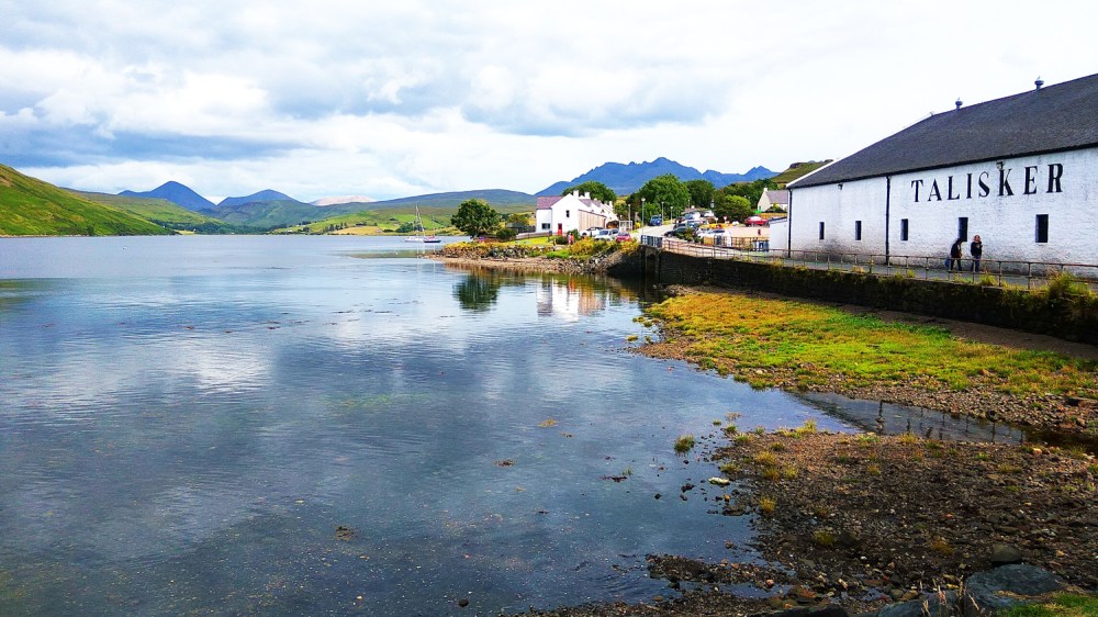 Destilería a orillas de un lago con montañas al fondo bajo un cielo nublado.