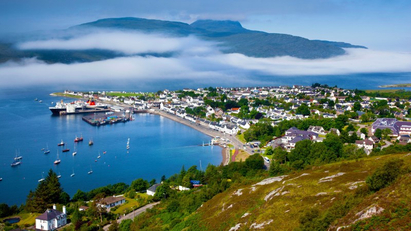 Coastal town with harbor, colorful houses, sailboats, and misty mountains in the background.