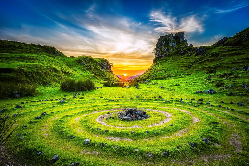 Bright sunset over lush green hills with stone spiral in foreground.