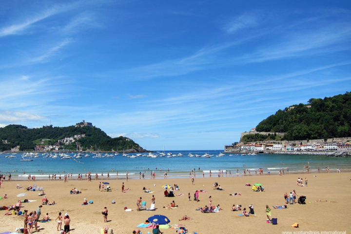 Crowded sandy beach with people sunbathing and swimming, green hills and boats on calm blue sea.