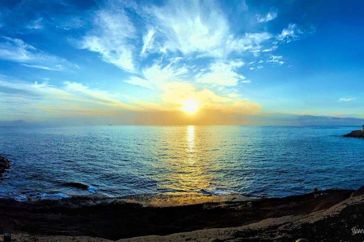 Panoramic view of a coastal sunset with vibrant blue sky and scattered clouds.