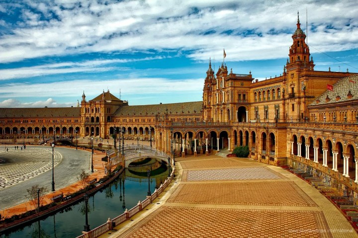 Wide view of Plaza de España, Seville, with curved building, arches, and a canal under blue sky.