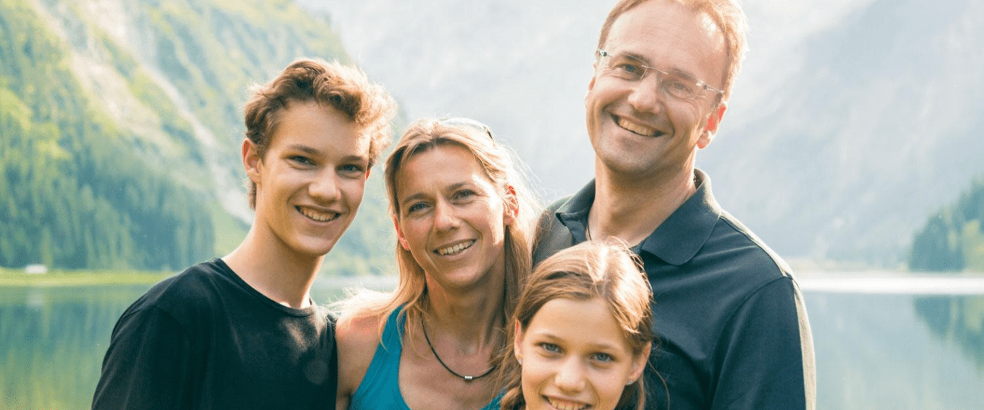 Family of four smiling by a lake with mountains in the background.
