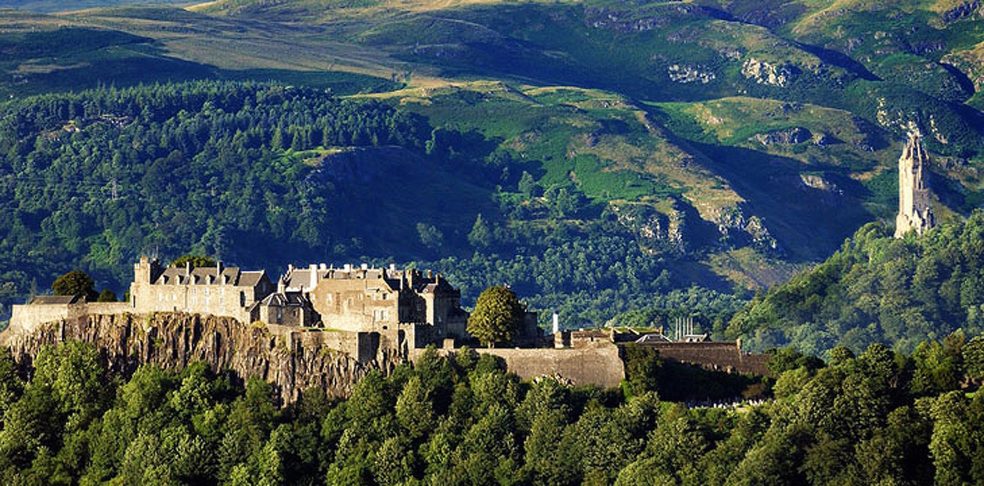 Castillo histórico de piedra enclavado en un acantilado, rodeado de exuberantes colinas verdes y árboles.