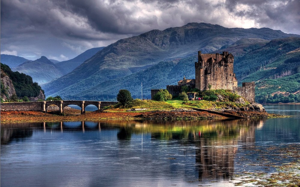 Castle on a small island with a stone bridge under cloudy skies near mountains and a calm lake.
